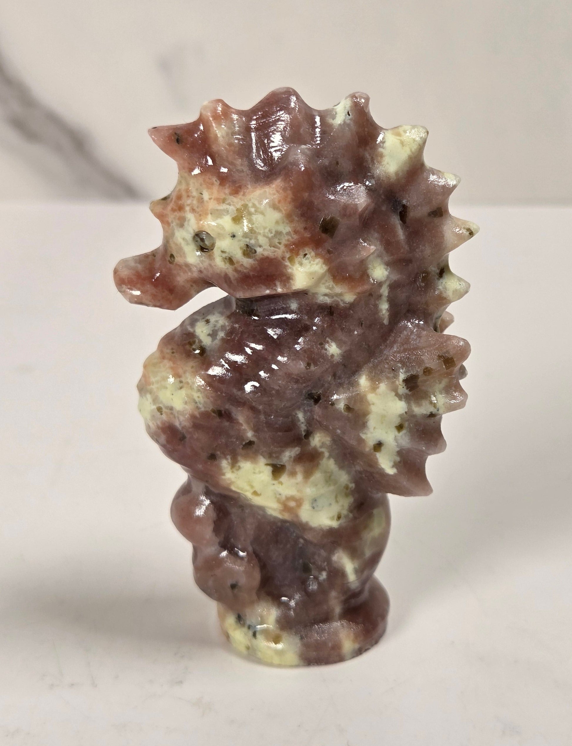 A small carved seahorse figurine made from polished stone with mottled brown, beige, and cream tones, standing on a light surface against a softly blurred marble-like background.