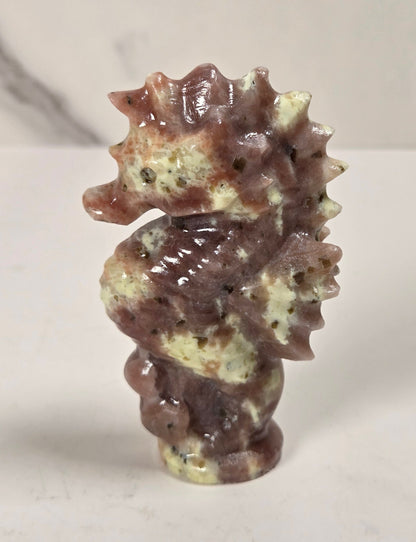 A small carved seahorse figurine made from polished stone with mottled brown, beige, and cream tones, standing on a light surface against a softly blurred marble-like background.