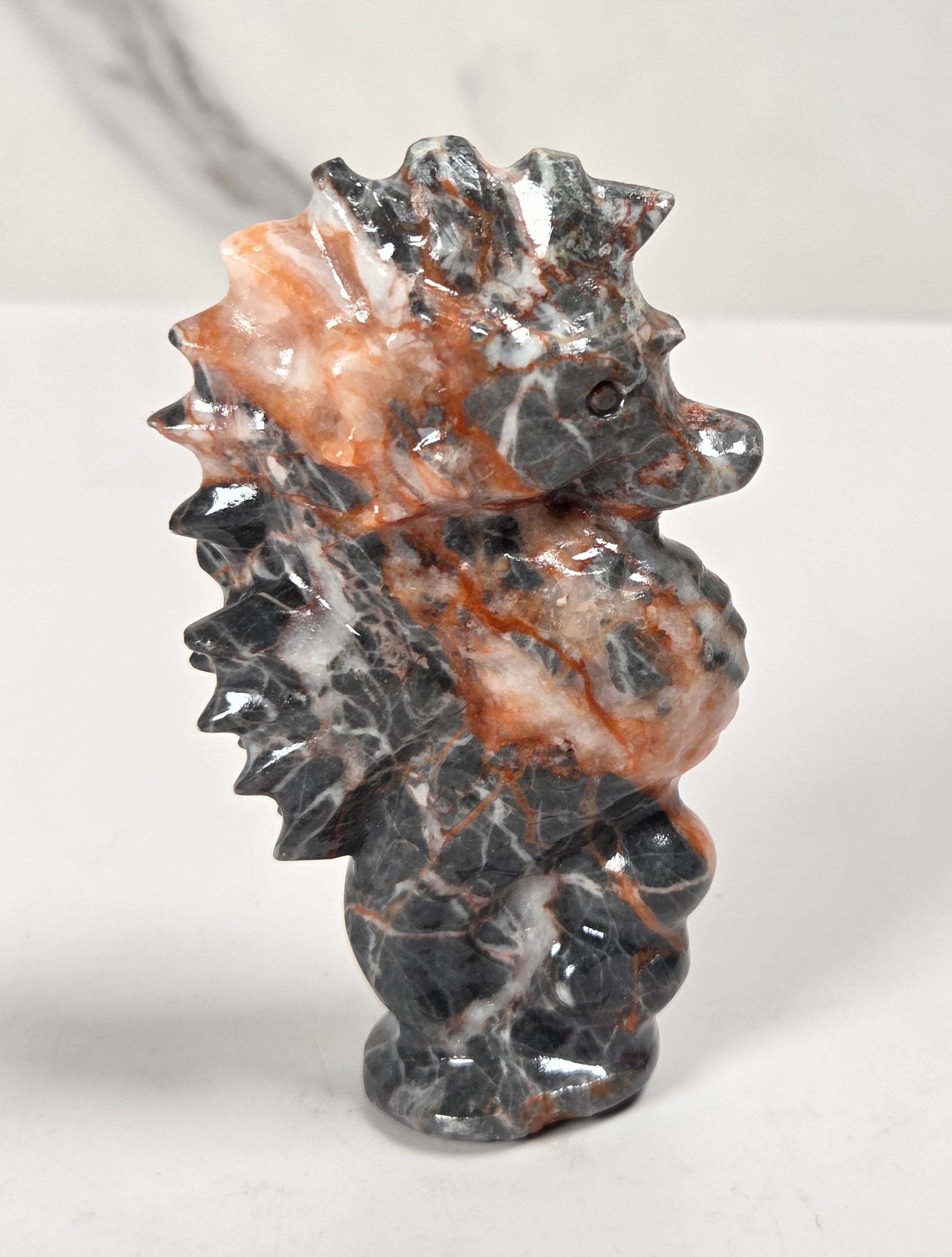 A small carved stone seahorse figurine with a glossy black and white marbled body and warm orange-brown veining, standing on a light surface against a pale marble-like background.