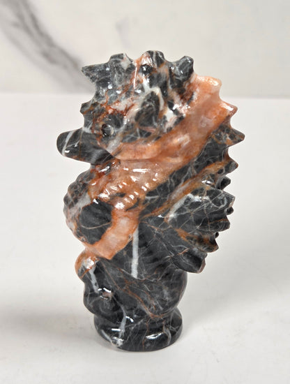 A small carved stone sculpture featuring a dark black and gray marbled body with a prominent copper-brown polished band running diagonally across the front, set against a light marble background.