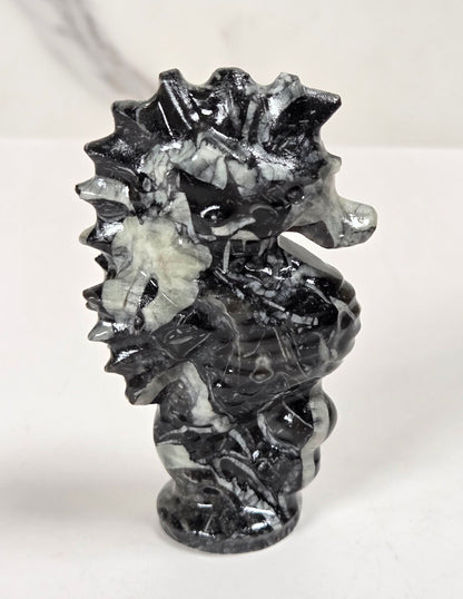 A small carved seahorse figurine made from black and white marbled stone, standing on a white surface against a light gray marble-patterned background.