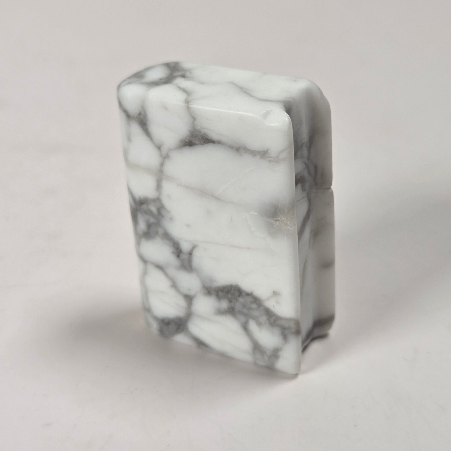 A rectangular lighter made of white marble with prominent gray veining, standing upright on a light gray surface.