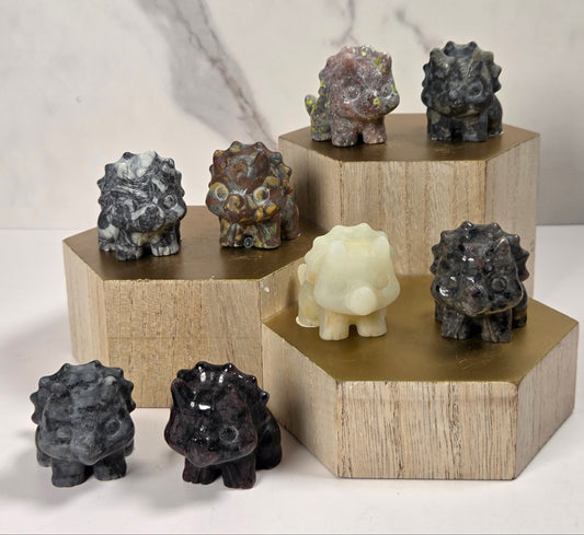 Seven small carved stone animal figurines displayed on hexagonal wooden pedestals against a light marble-look background. The figurines are made from various natural stones with different colors, including black, gray, brown, cream, and marbled patterns.