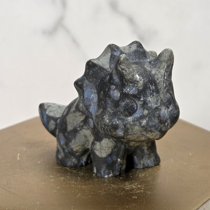 A small dark gray and black spotted dinosaur figurine, resembling a Triceratops, displayed on a hexagonal gold-colored pedestal against a light marble background.