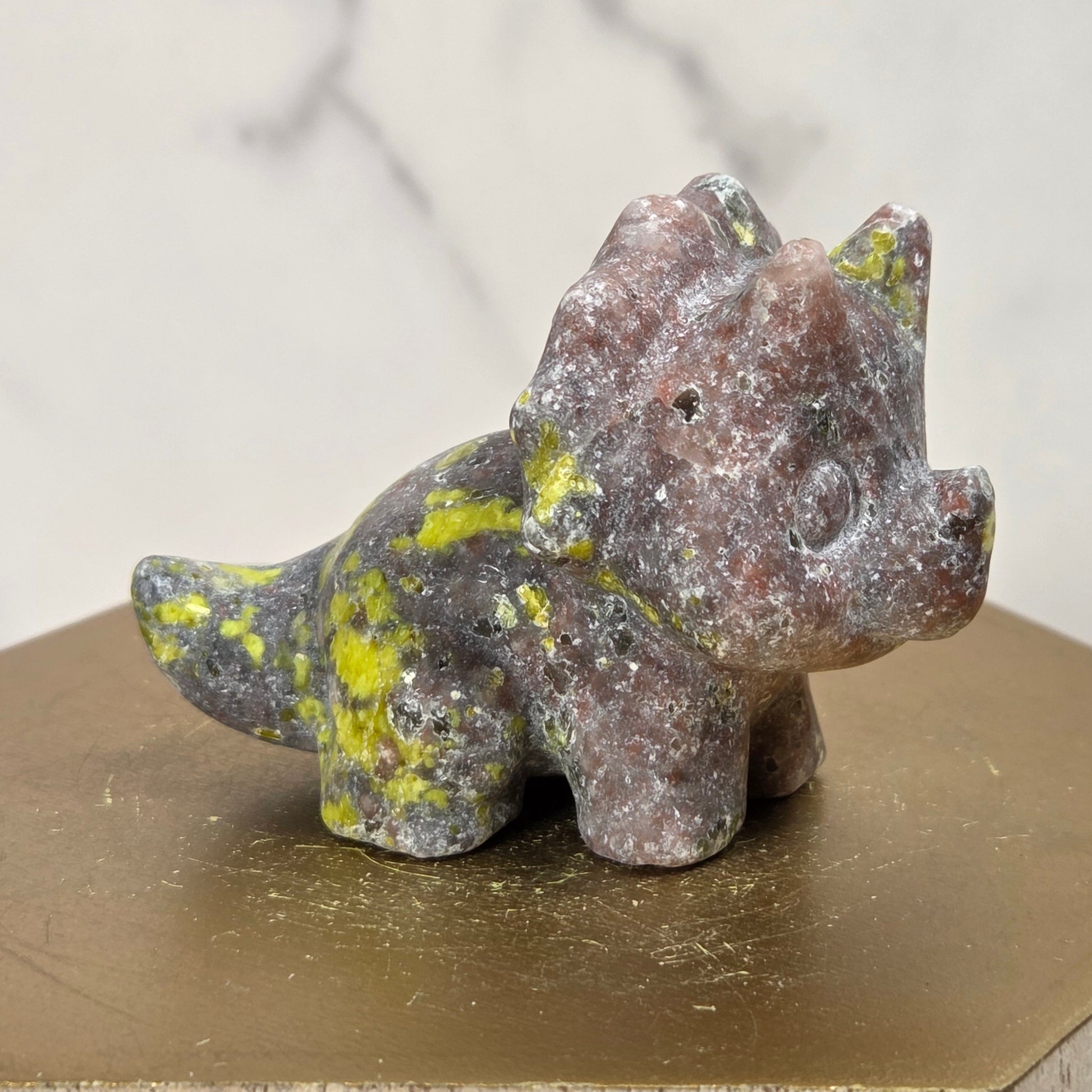 A small natural stone triceratops figurine with a reddish-brown and gray marbled texture and bright yellow-green veining, displayed on a gold hexagonal pedestal.