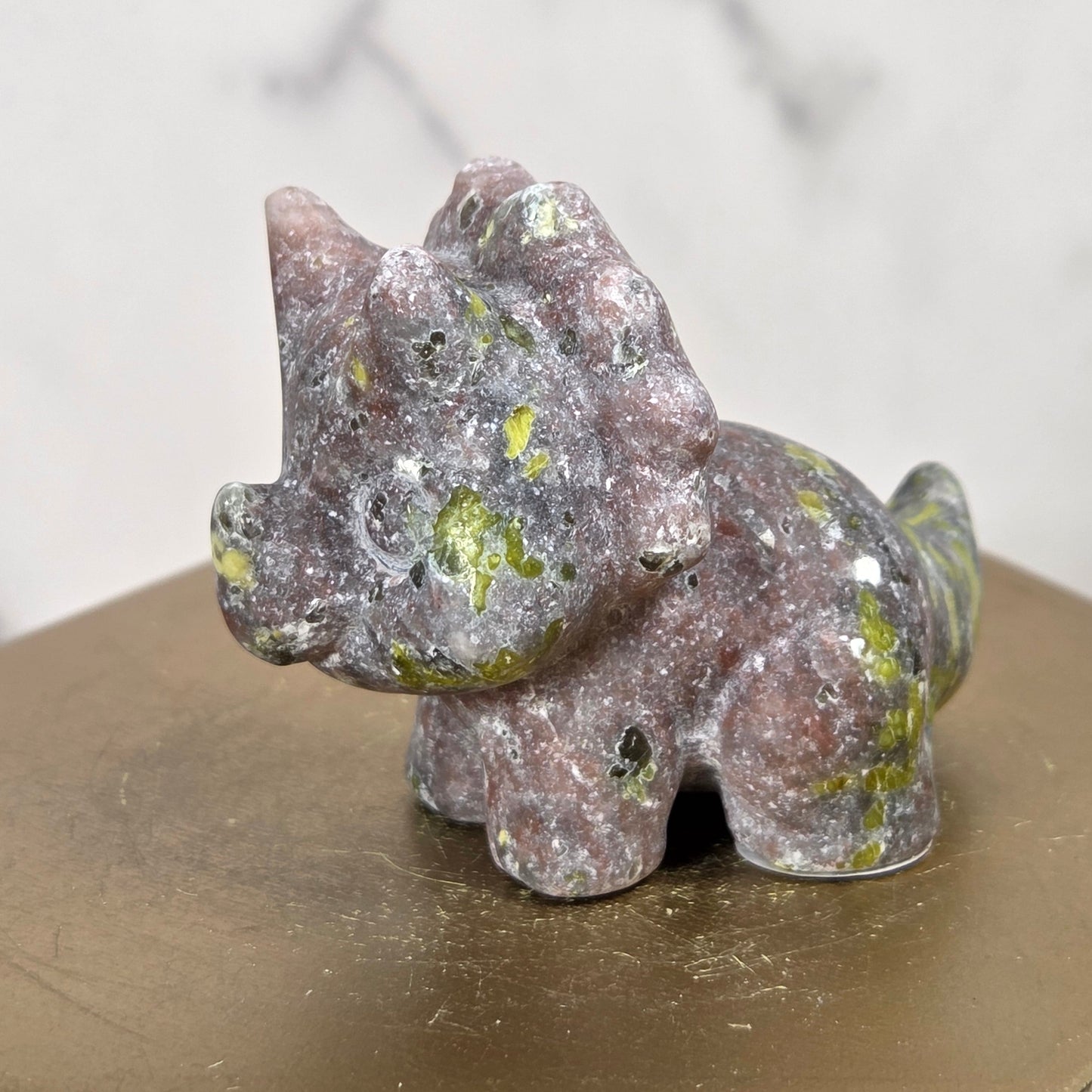 A small carved stone figurine of a horned animal, likely a deer or antelope, with a mottled gray, pinkish, and green natural stone texture, displayed on a hexagonal gold-toned pedestal.