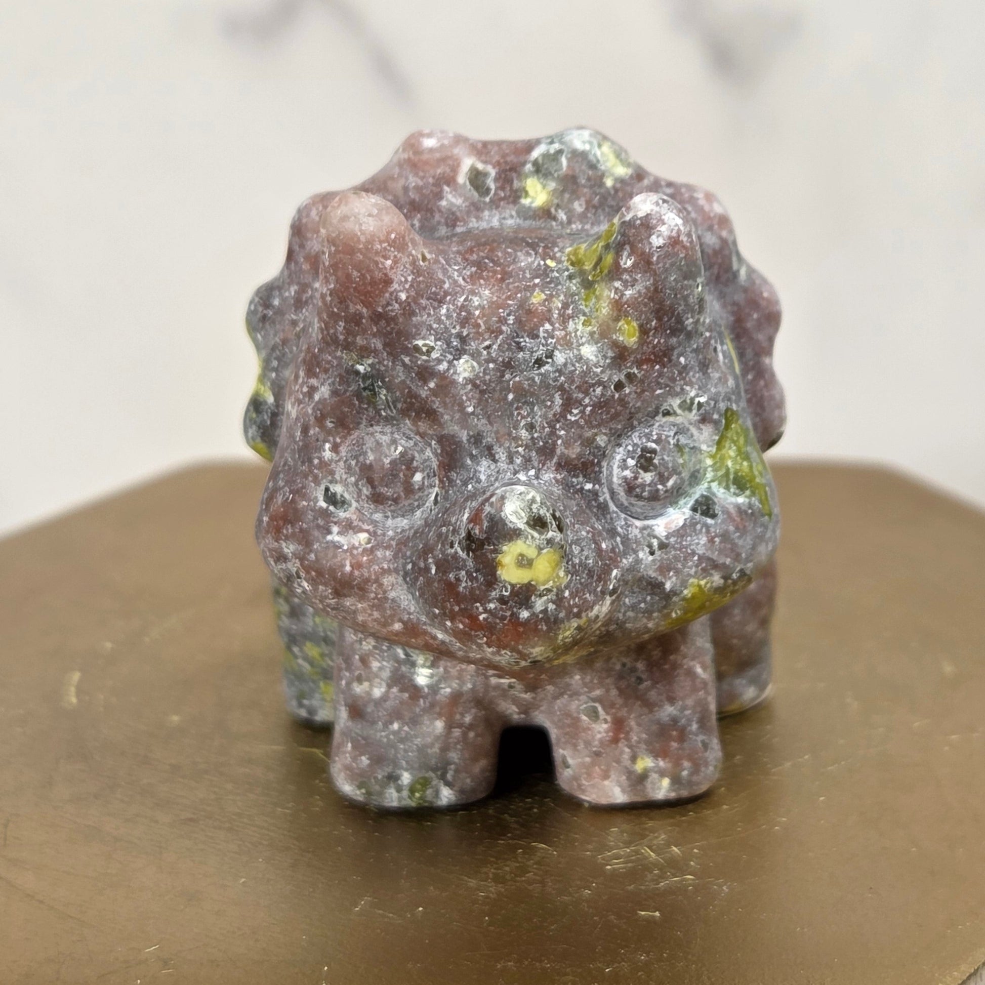 A small carved stone figurine with a rough, textured surface in earthy brown, gray, and green tones, displayed on a gold-colored hexagonal block.