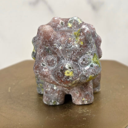 A small carved stone figurine with a rough, textured surface in earthy brown, gray, and green tones, displayed on a gold-colored hexagonal block.
