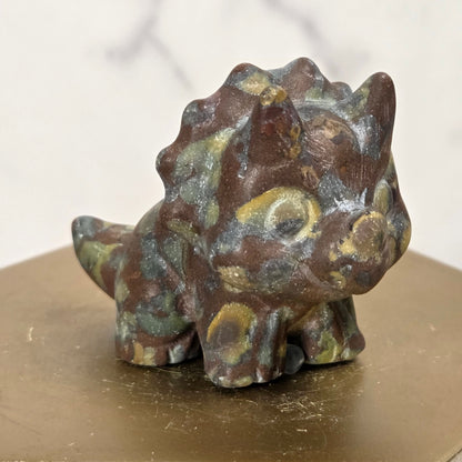 A small decorative dinosaur figurine with a textured, patinated surface in brown, green, and gold tones, displayed on a hexagonal gold-toned pedestal against a light marble-patterned background.