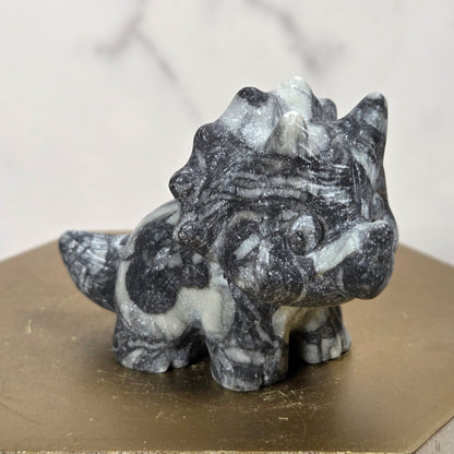 A small carved dinosaur figurine made of black and white marbled stone, displayed on a hexagonal gold-colored block against a light marble-patterned background.