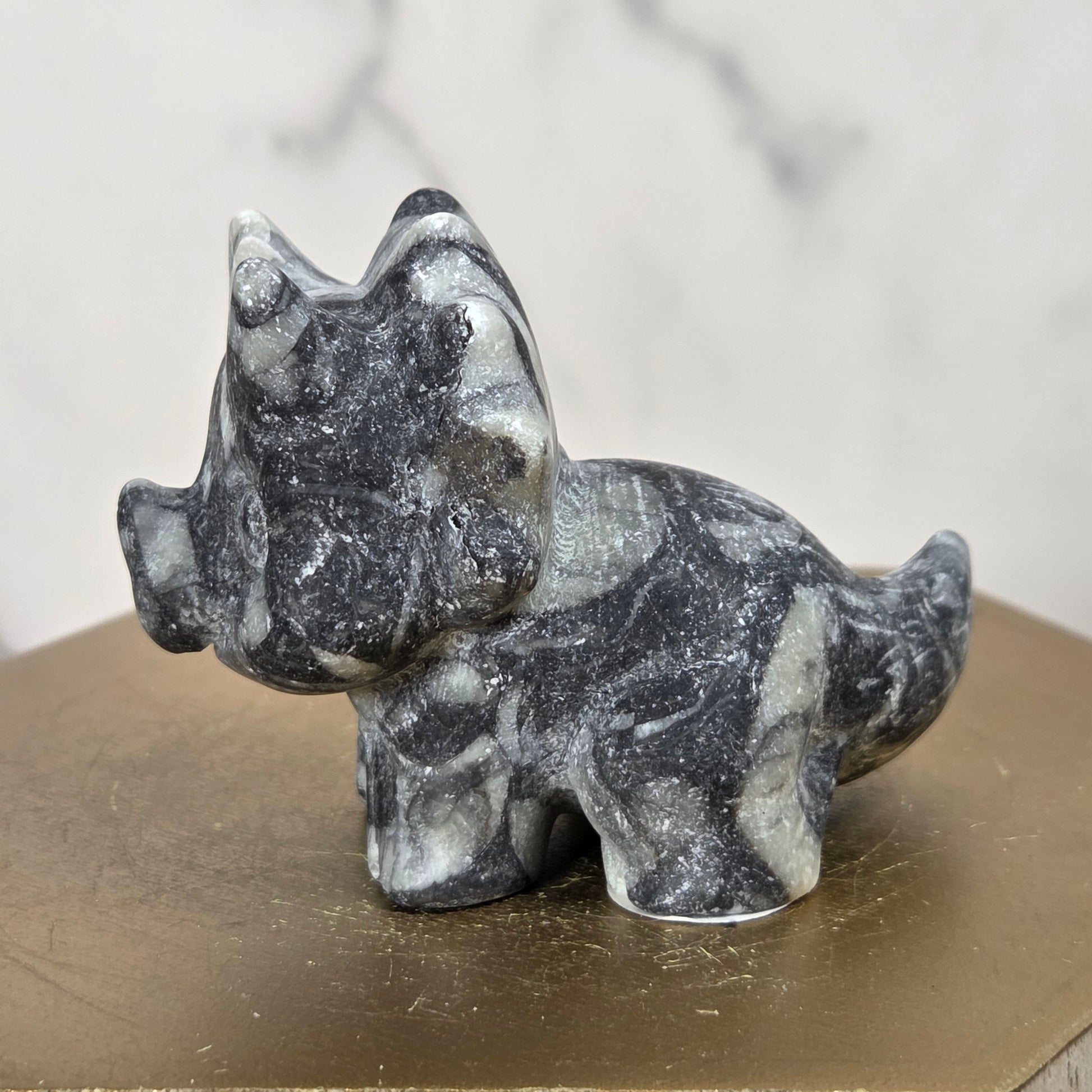 A small carved stone dinosaur figurine, made from black and white marbled stone, displayed on a hexagonal gold-toned wooden block against a light marble background.