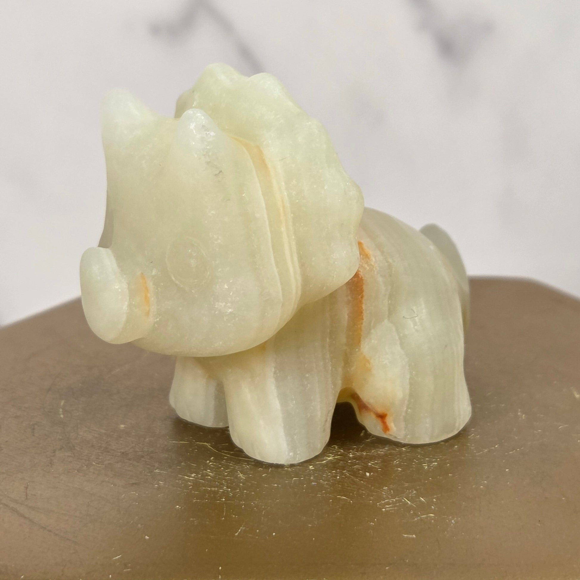 A small carved animal figurine, shaped like a dinosaur, made from pale cream-colored stone with subtle orange-brown veining. The figurine is displayed on a hexagonal wooden block against a light gray marble-patterned background.
