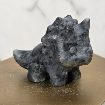 A small dark gray stone dinosaur figurine, resembling a Triceratops, displayed on a gold-colored hexagonal block pedestal against a light marble background.