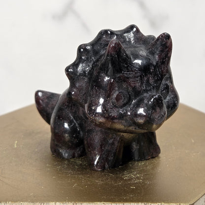 A small glossy black stone dinosaur figurine, resembling a Triceratops, displayed on a gold-colored hexagonal pedestal against a light marble background.