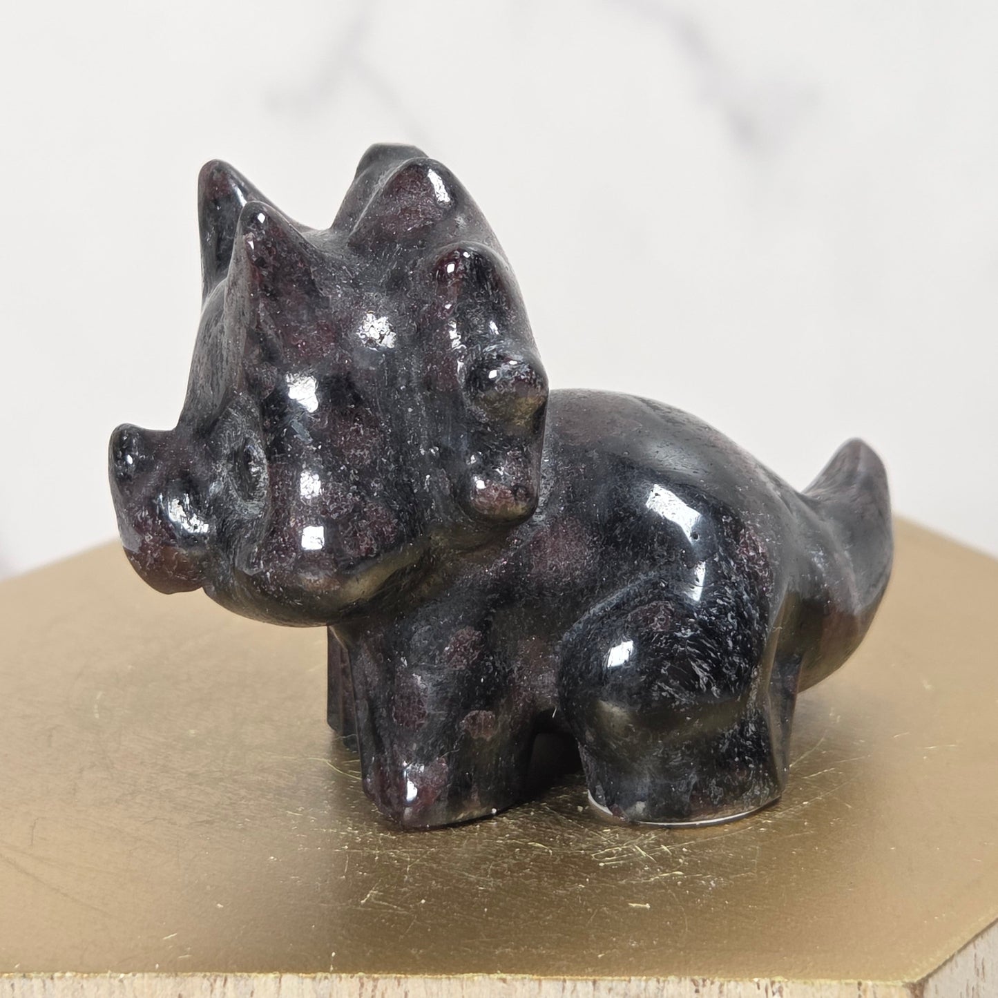 A small glossy black stone figurine of a horned animal, likely a stag or deer, displayed on a gold-colored hexagonal block against a light marble background.