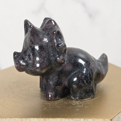 A small glossy black stone figurine of a horned animal, likely a stag or deer, displayed on a gold-colored hexagonal block against a light marble background.