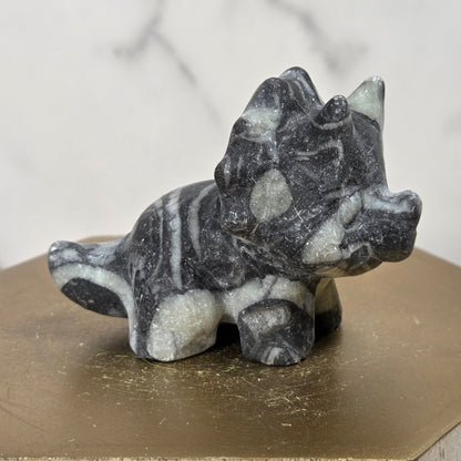 A small carved stone triceratops figurine in dark gray and white marbled stone, displayed on a hexagonal gold-toned pedestal against a light marble-patterned background.