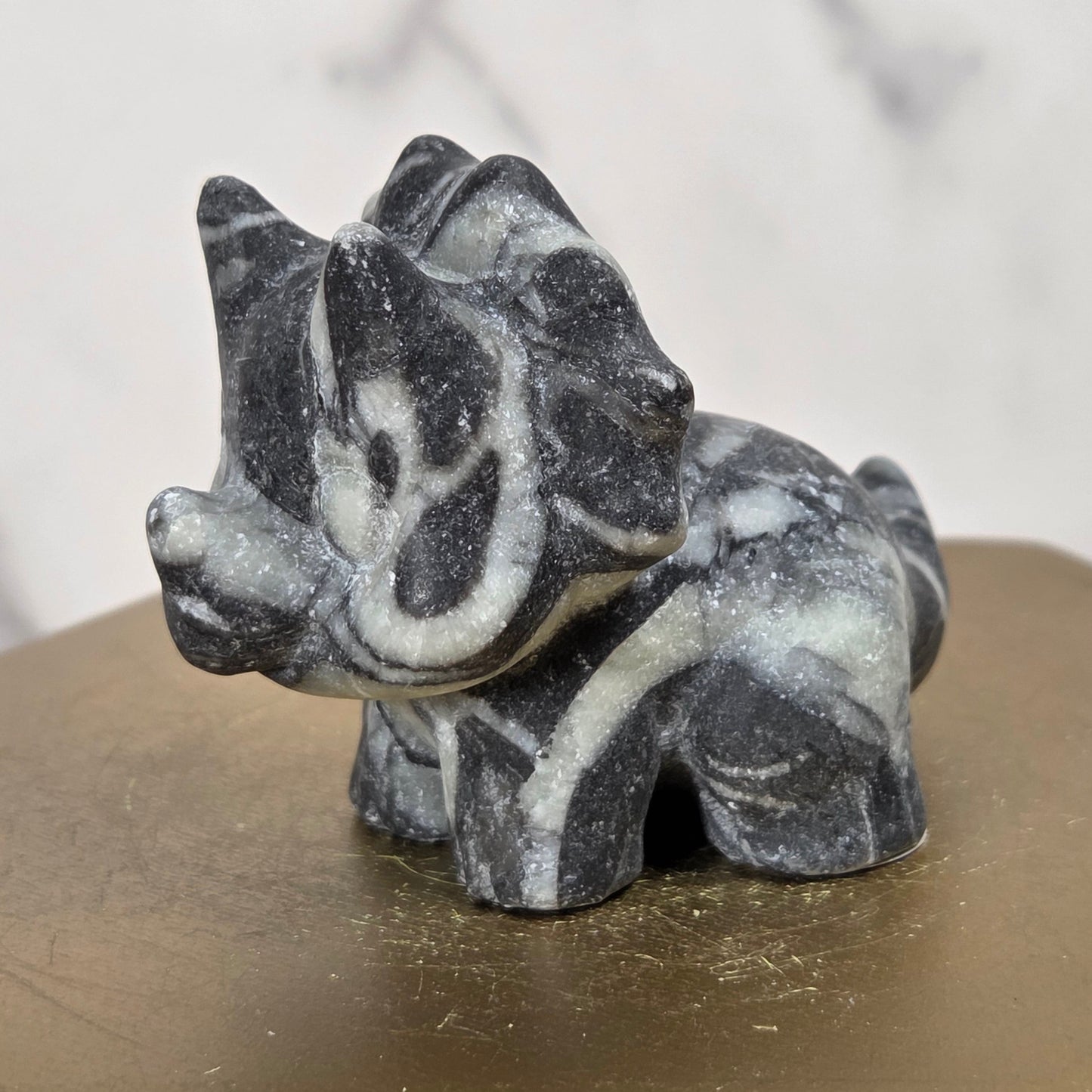 A small carved animal figurine, likely a deer or antelope, made from dark and light marbled stone, displayed on a hexagonal gold-colored pedestal.