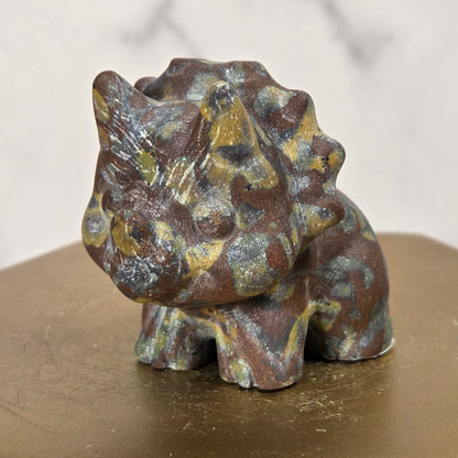 A small decorative dragon figurine with a textured, weathered finish in brown, bronze, and muted green, displayed on a hexagonal gold-toned pedestal.