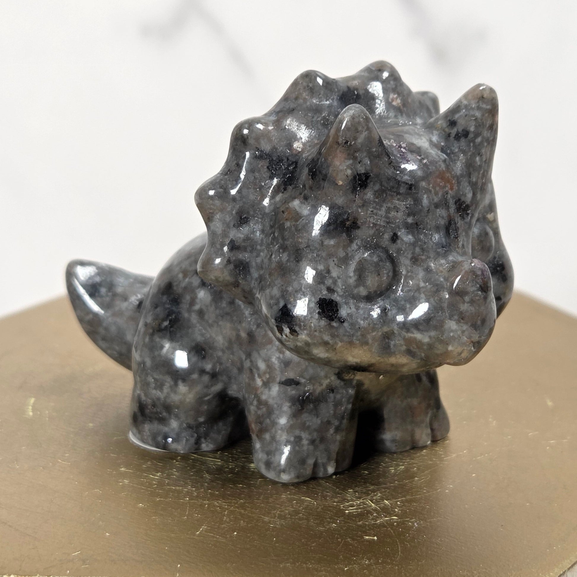 A small polished dark gray stone dinosaur figurine, resembling a Triceratops, displayed on a gold-colored hexagonal block against a light marble background.