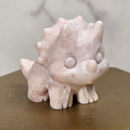 A small pale pink dinosaur figurine with a textured, stone-like finish, standing on a hexagonal metallic gold pedestal against a soft gray marble background.