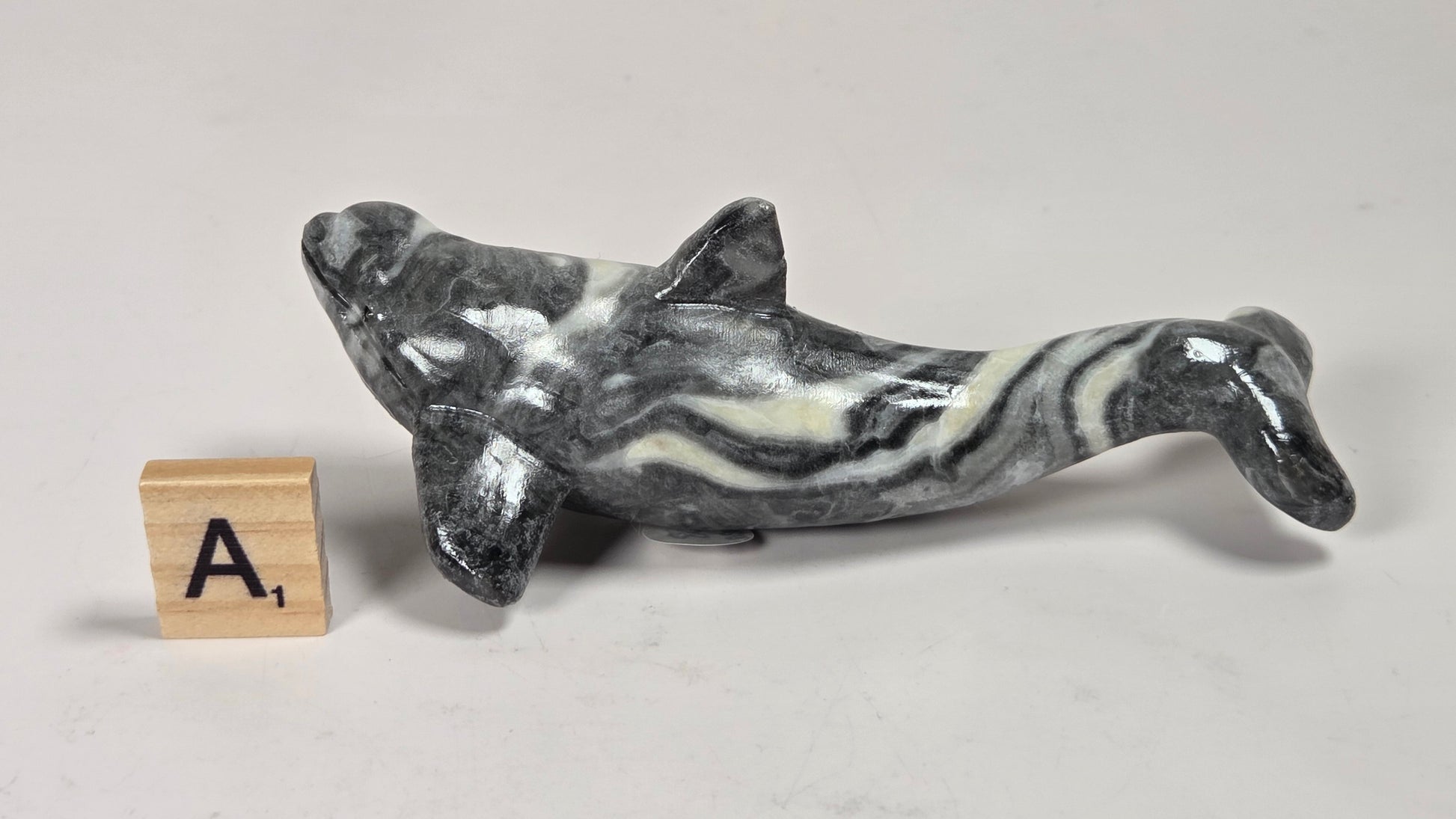 A small polished stone dolphin figurine with a black, white, and gray marbled pattern, shown next to a wooden letter block displaying the letter 'A' on a light gray surface.