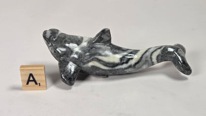 A small polished stone dolphin figurine with a black, white, and gray marbled pattern, shown next to a wooden letter block displaying the letter 'A' on a light gray surface.