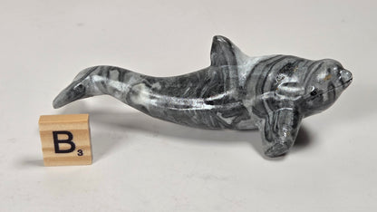 A polished gray and white marbled dolphin figurine shown on a light gray background next to a wooden Scrabble tile with the letter B.