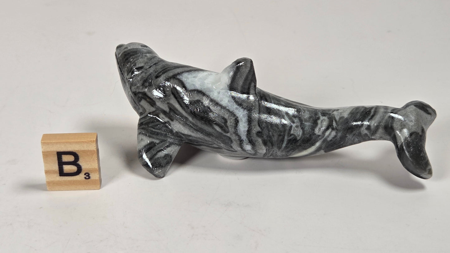 A polished black and white marbled dolphin figurine displayed on a light gray surface next to a wooden letter 'B' tile.