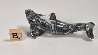 A polished black and white marbled dolphin figurine displayed on a light gray surface next to a wooden letter 'B' tile.