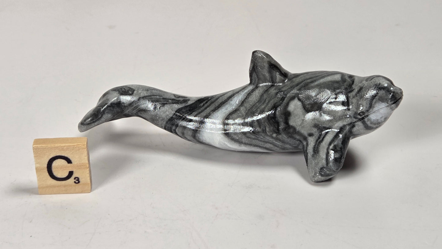 A small polished whale or dolphin figurine with a black, white, and gray marbled pattern, shown on a light gray background next to a wooden Scrabble tile showing the letter C and value 3.