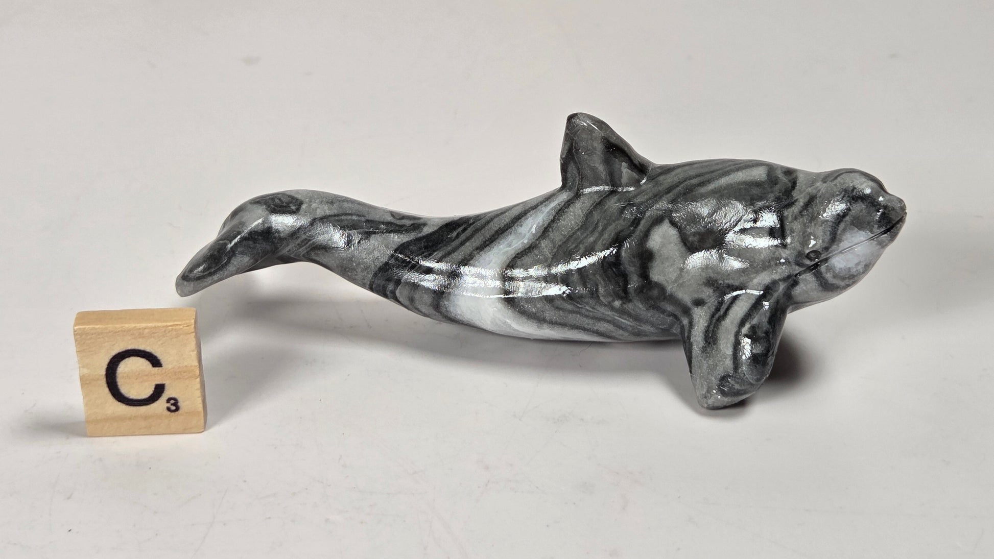 A small polished whale or dolphin figurine with a black, white, and gray marbled pattern, shown on a light gray background next to a wooden Scrabble tile showing the letter C and value 3.