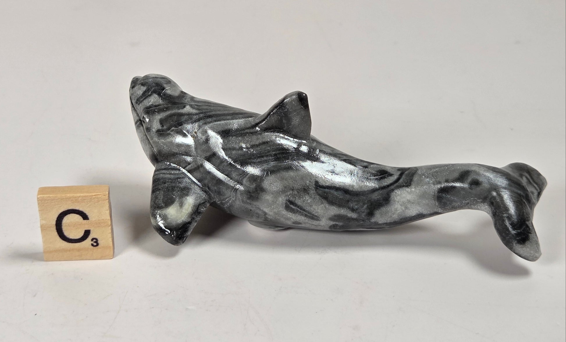 A small polished gray and black marbled dolphin figurine displayed on a light gray surface next to a wooden Scrabble tile showing the letter C.
