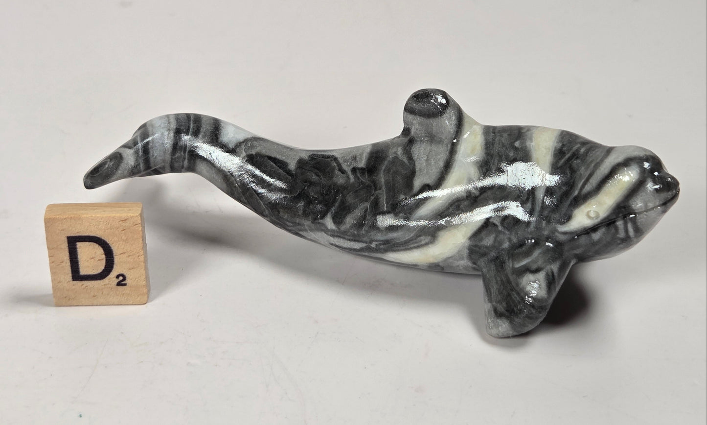 A small sculpted dolphin figurine with a black, white, and gray marbled pattern, shown next to a wooden Scrabble tile with the letter D2 on a light gray background.