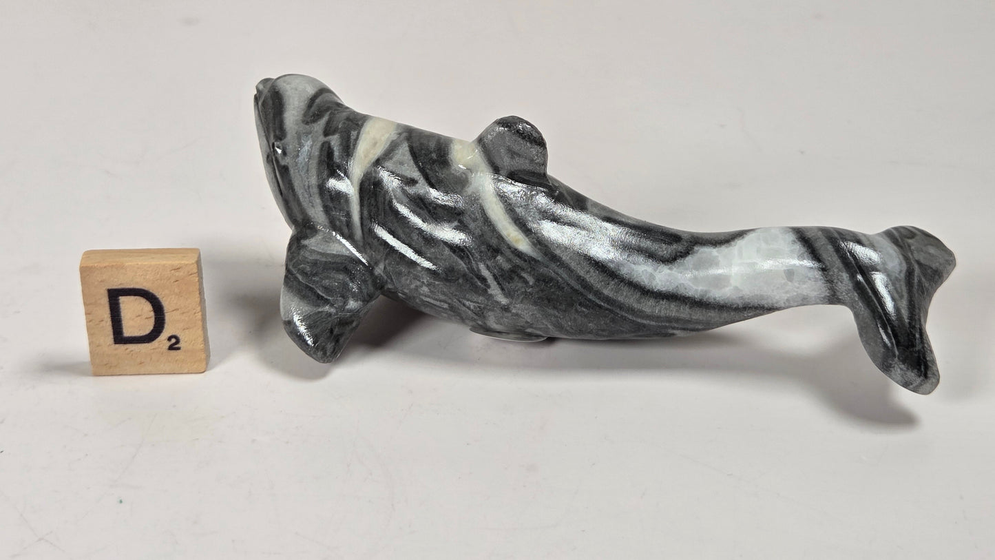 A small carved figurine of a fish, made from black and white marbled stone, displayed next to a wooden Scrabble tile on a plain light gray surface.
