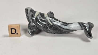 A small carved figurine of a fish, made from black and white marbled stone, displayed next to a wooden Scrabble tile on a plain light gray surface.