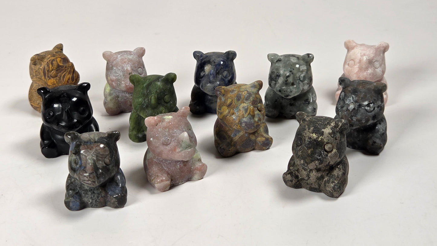 A group of small carved rhinocerus  figurines, made from natural stone with varied earthy colors like black, green, brown, pink, and gray, arranged on a plain white surface.