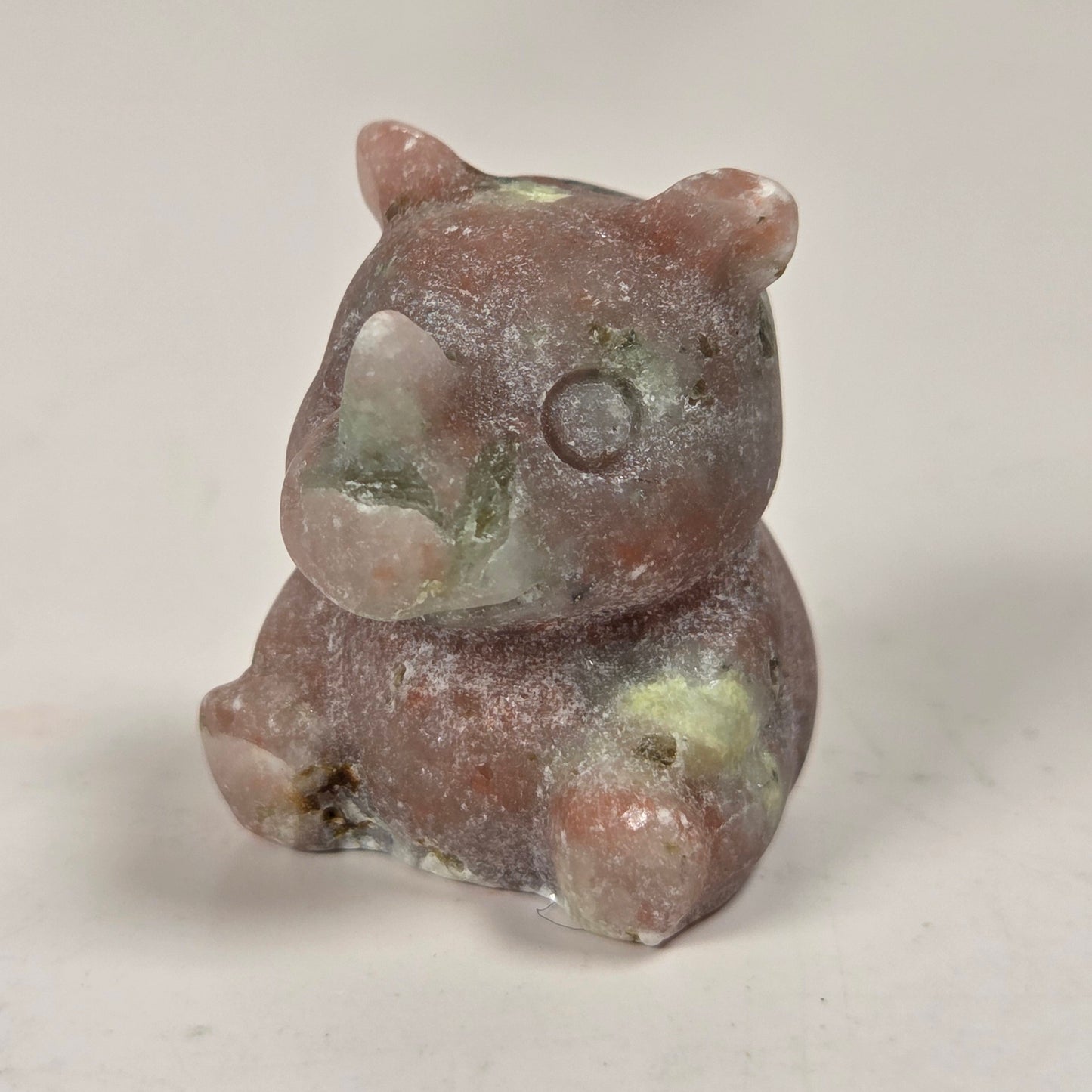 A small carved figurine of a bear made from a natural stone, featuring a dusty, textured finish with earthy brown, pink, and green hues, set against a plain light background.
