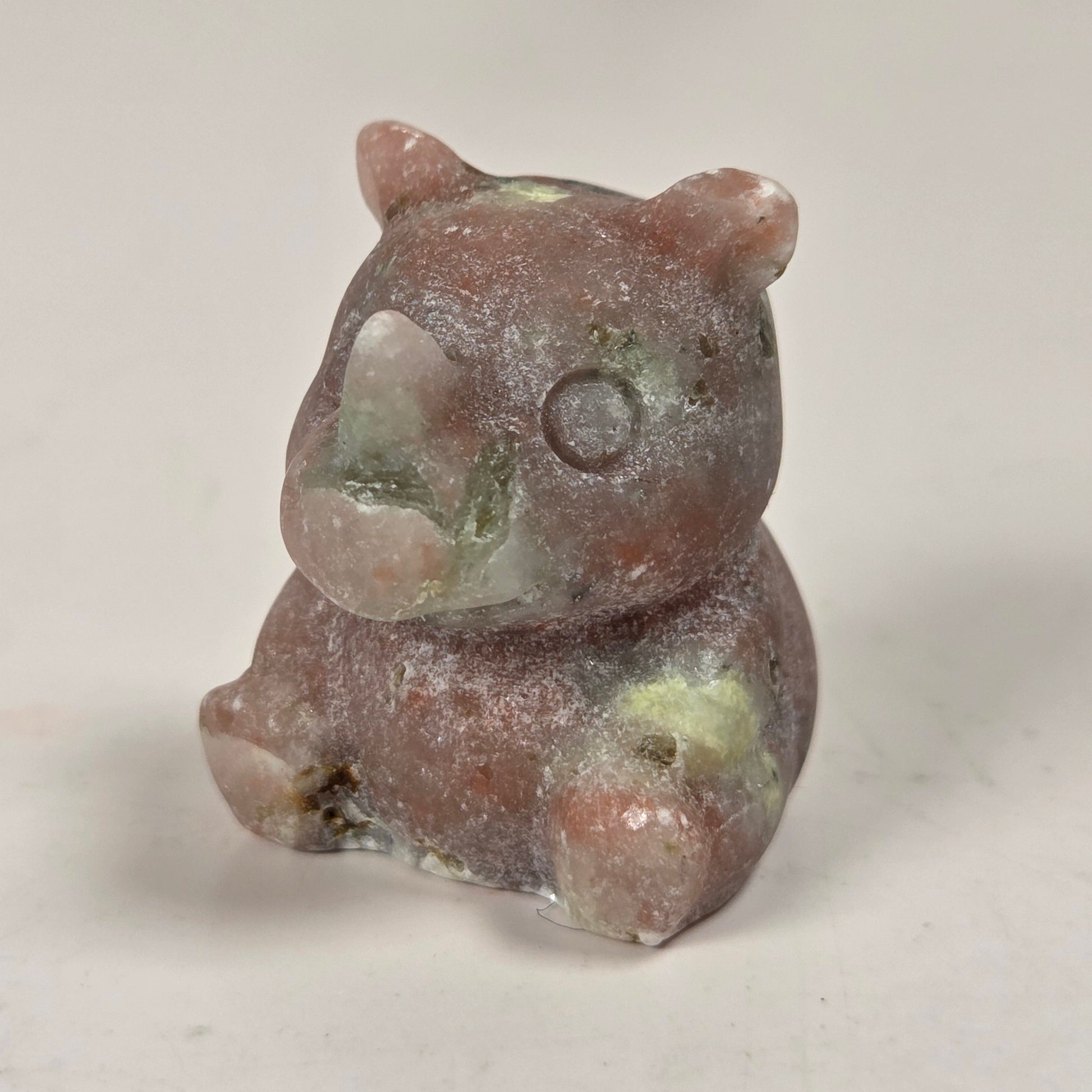 A small carved figurine of a bear made from a natural stone, featuring a dusty, textured finish with earthy brown, pink, and green hues, set against a plain light background.