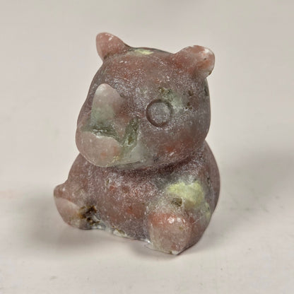 A small carved figurine of a bear made from a natural stone, featuring a dusty, textured finish with earthy brown, pink, and green hues, set against a plain light background.