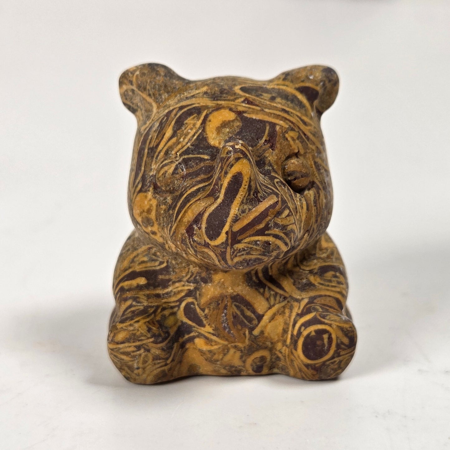 A small carved figurine of a bear sitting cross-legged, made from a marbled wood or stone with swirling brown and golden-yellow patterns, set against a plain off-white background.