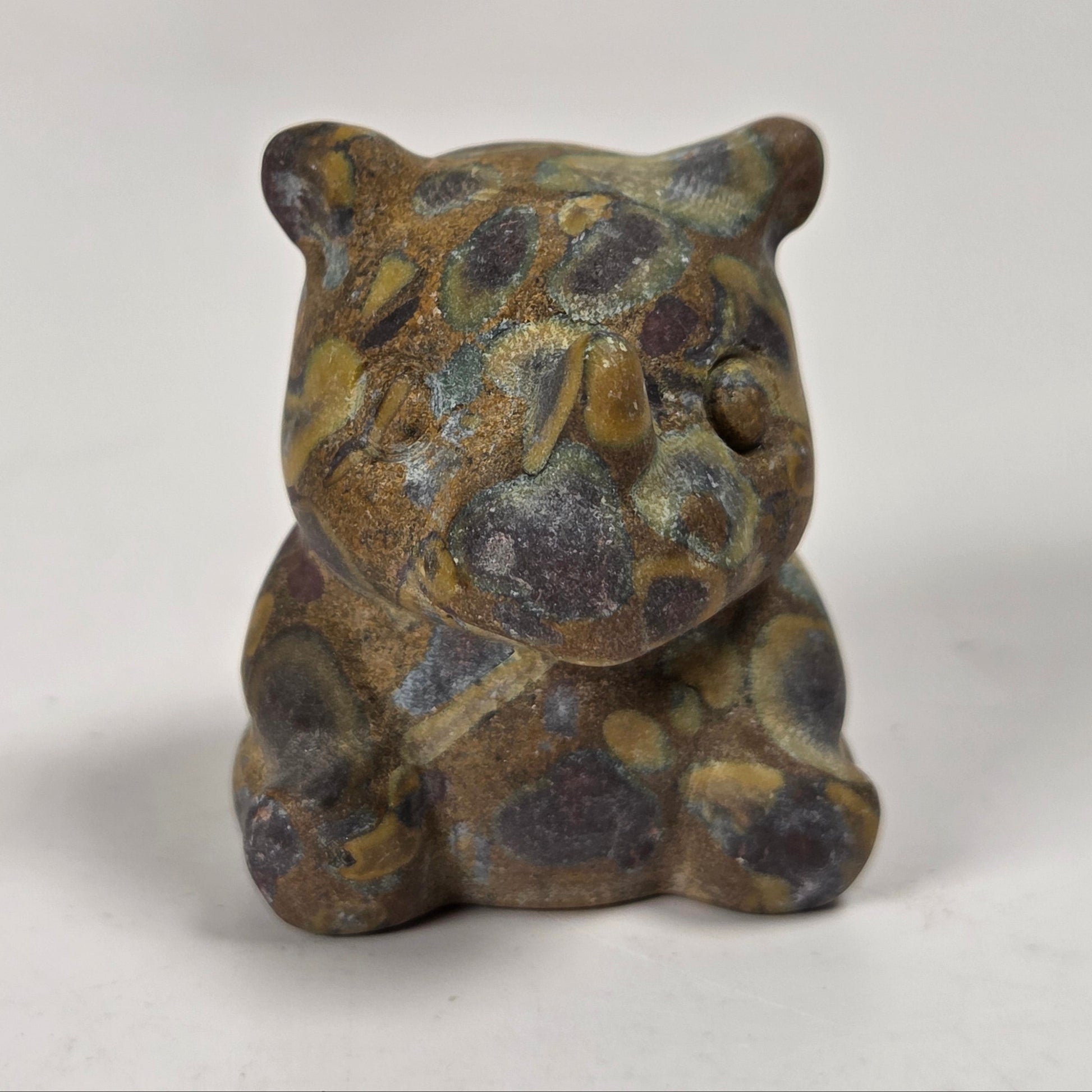 A small sculpted figurine of a seated animal, likely a bear or cat, with a mottled stone-like appearance in earthy brown, gray, and muted gold tones, photographed against a plain light background.
