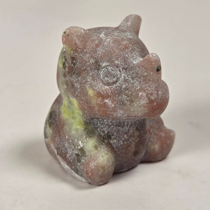 A small carved figurine of a seated animal, likely a pig or boar, made from a pinkish stone with green and gray marbling, photographed on a plain light background.