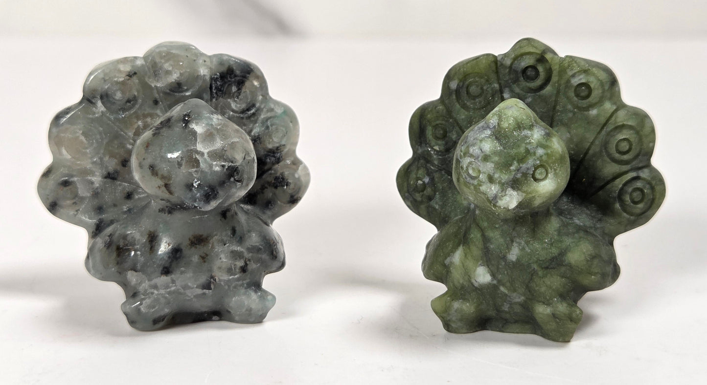 Two small carved stone figurines, likely peacock or toad-like forms, displayed side by side on a white surface with a light marble-look background. One is gray with black speckling, and the other is olive green with white marbling.