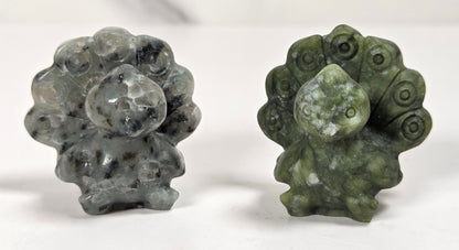 Two small carved stone figurines, likely peacock or toad-like forms, displayed side by side on a white surface with a light marble-look background. One is gray with black speckling, and the other is olive green with white marbling.