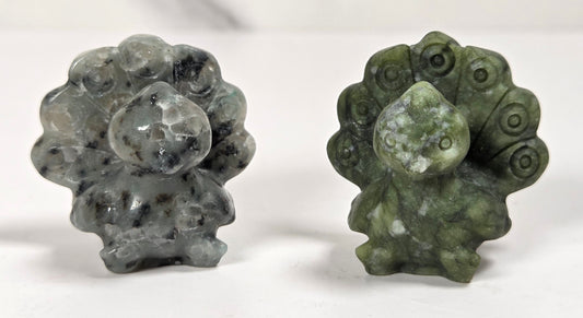 Two small carved stone figurines, likely peacock or toad-like forms, displayed side by side on a white surface with a light marble-look background. One is gray with black speckling, and the other is olive green with white marbling.