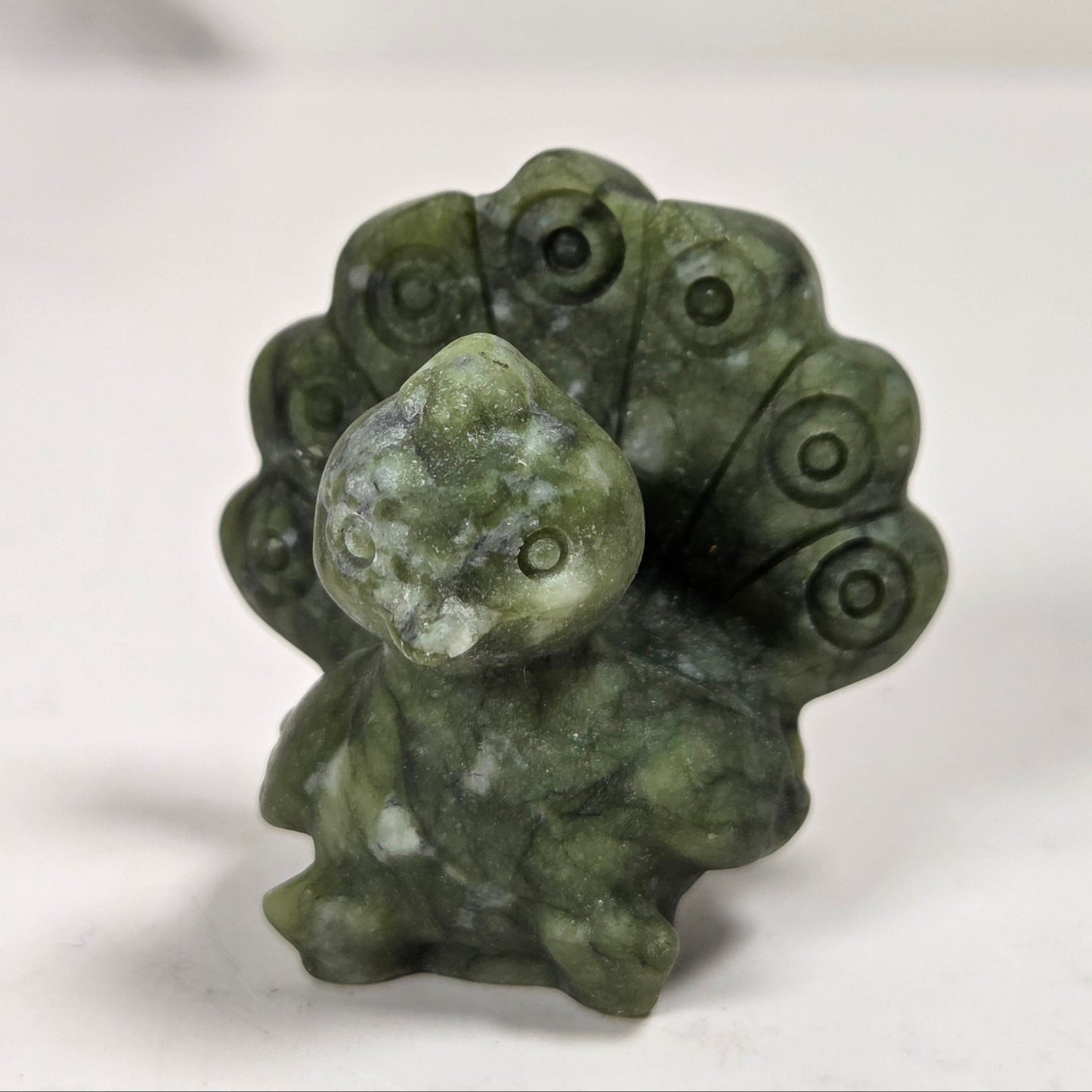 A small carved green stone figurine of a peacock with its tail feathers fanned out, standing on a light gray surface.