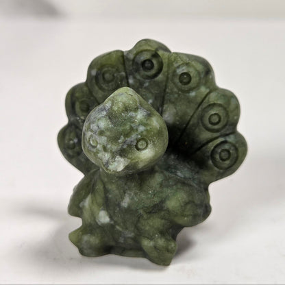 A small carved green stone figurine of a peacock with its tail feathers fanned out, standing on a light gray surface.