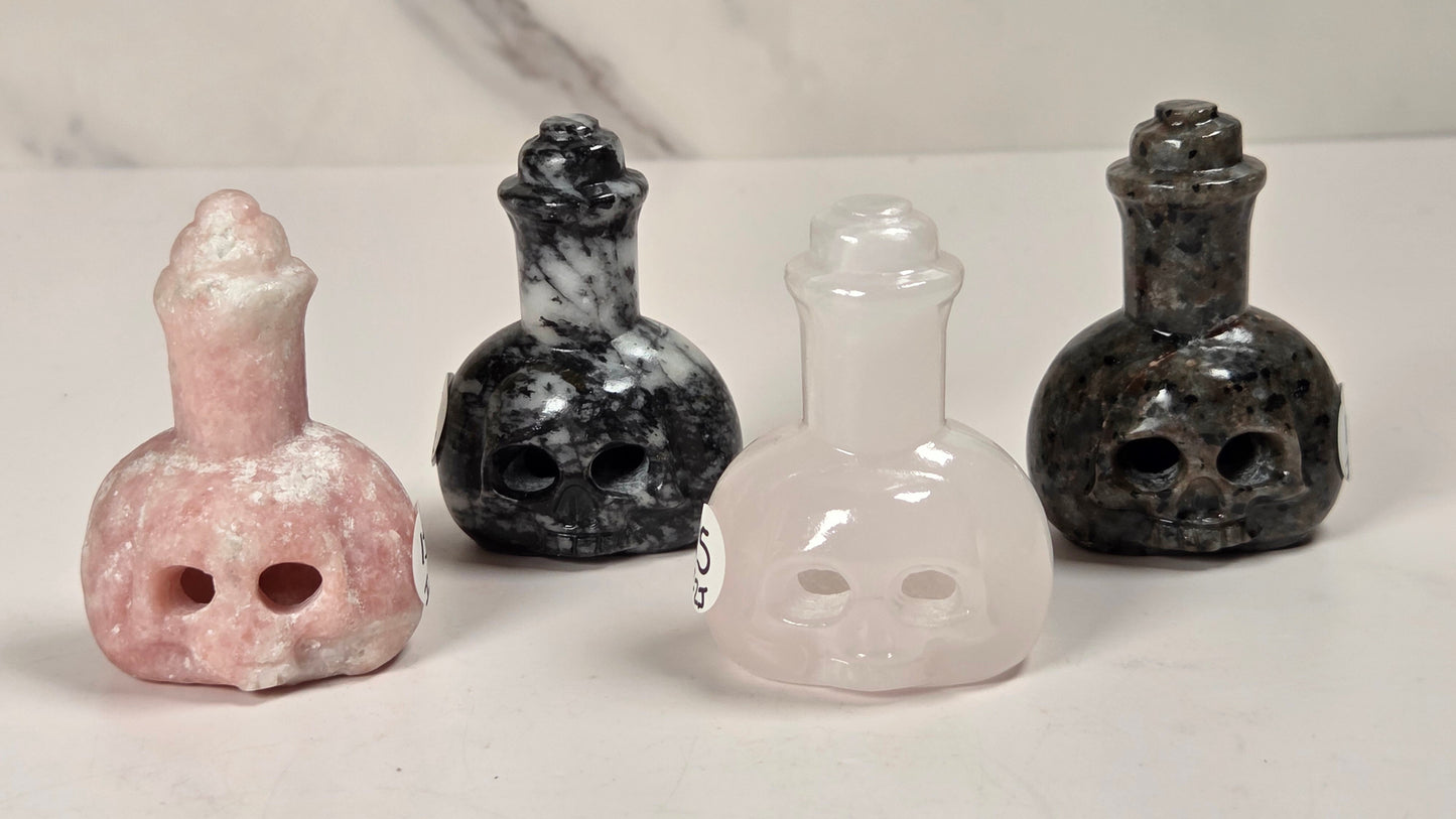 Four small skull-shaped bottle carvings made from different stones, arranged in a row on a light surface. From left to right: pink stone, black and white marbled stone, pale pink stone, and dark gray/black marbled stone.