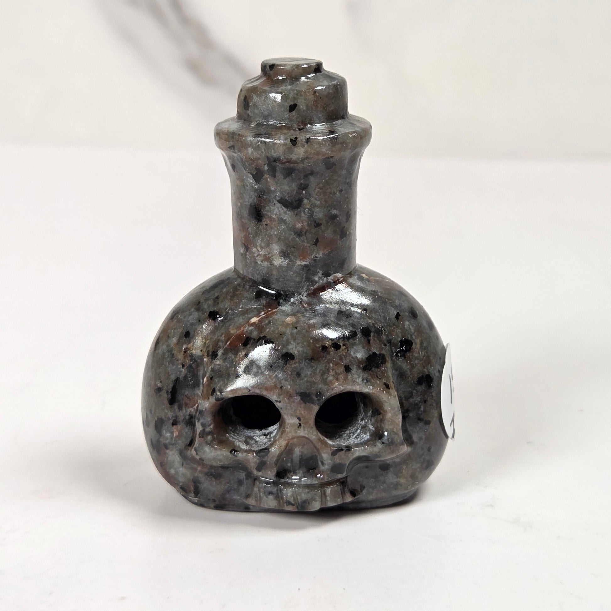 A small skull-shaped bottle or pendant made of polished dark stone with black, gray, and brown speckling, shown on a plain light background.